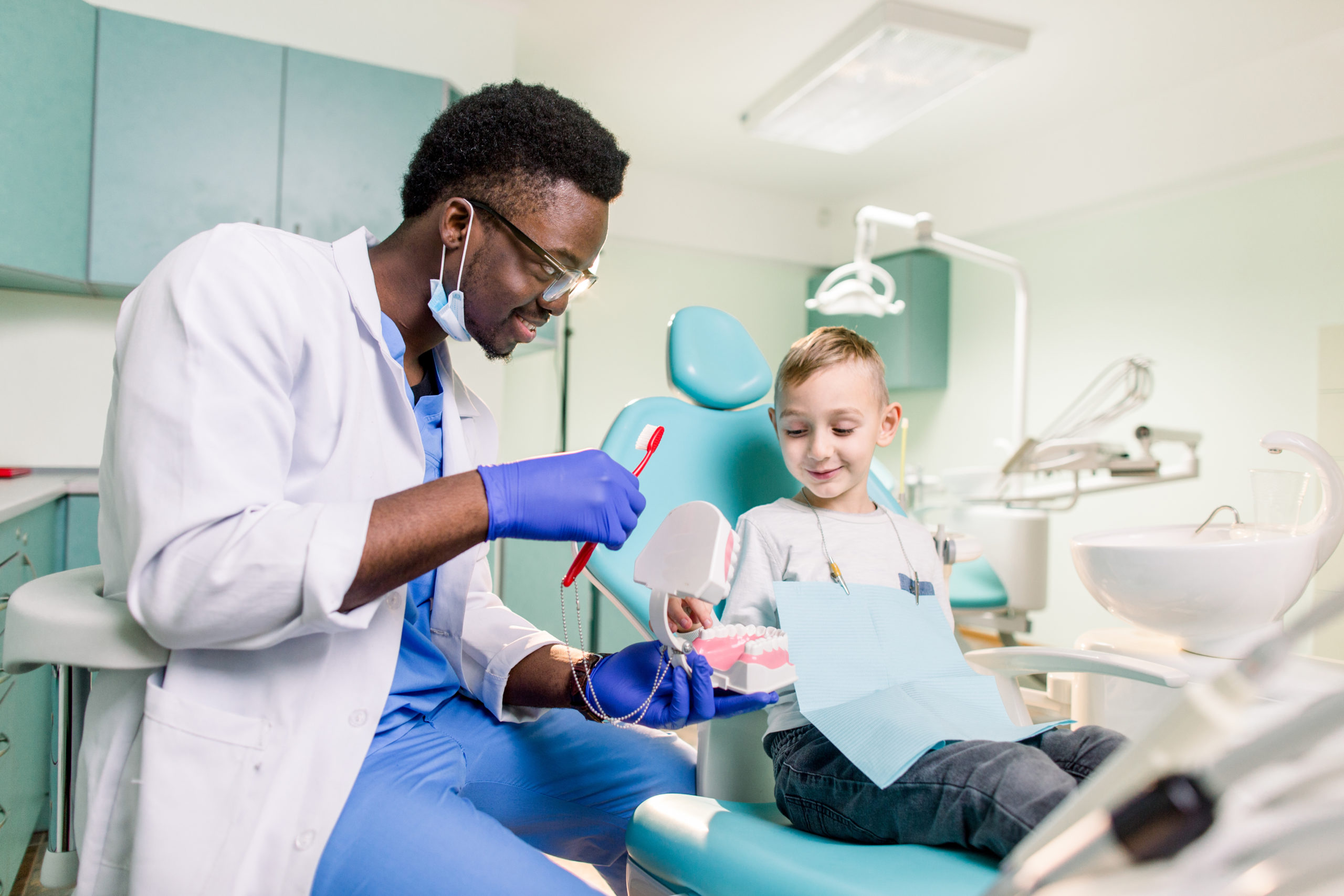 Happy smiling black dentist tells Caucasian little boy how to brush her teeth. Caries prevention, Dentistry, teeth hygiene concept